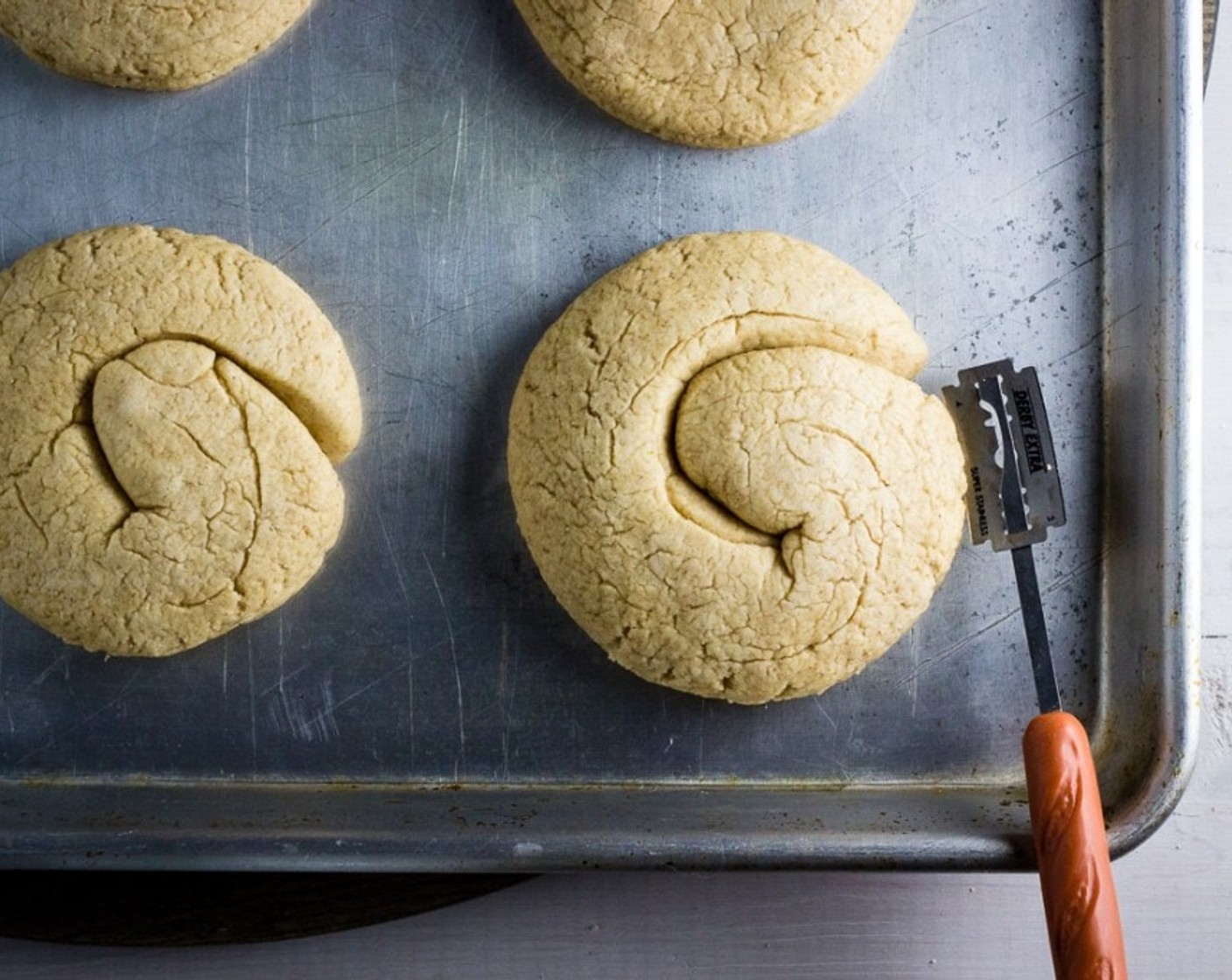 step 8 Place paximadia onto baking sheets about 2 inches apart (6 per baking sheet). Score each paximadi around the edges with a lame or a sharp knife to facilitate easier slicing in half once baked. Cover paximadia and let rise for 1 hour.