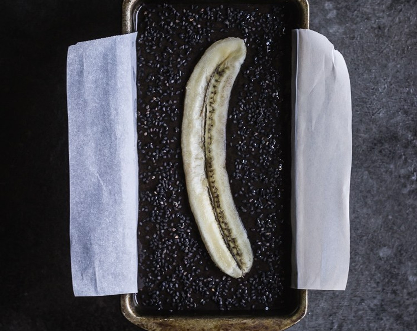 step 7 Lightly grease a 9x5-inch metal baking pan & line it with a sheet of parchment paper. Pour the batter into the baking pan, top with remaining Toasted Black Sesame Seeds (1 tsp), the sliced banana and then sprinkle the remaining sugar.