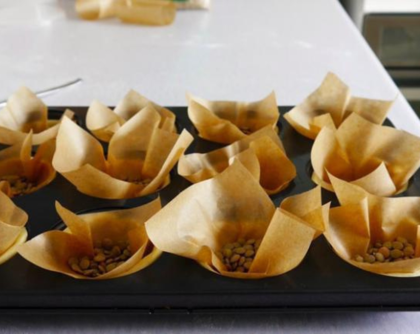 step 2 Place each piece in a muffin tin with no oil or butter. Press gently to make the dough stick to the side of the tin as to create a well formed whole in the middle and with a fork prick 2 or 3 times each circle to allow steam to flow whilst baking. Once all the dough circles are done place squares pieces of parchment or baking paper over each tart holding it down with some baking beans or rice, lentils, beans