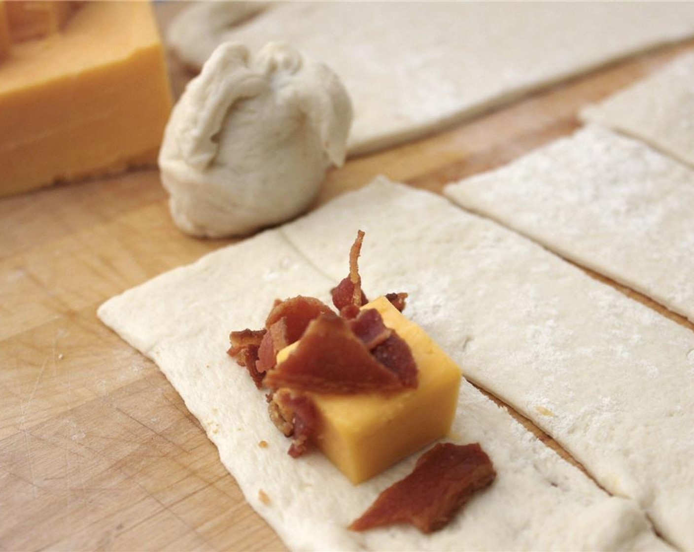 step 2 Cut the Pizza Dough (1 pckg) into approx 3x5-inch strips. Place a cube of Cheddar Cheese (2 cups) and some Bacon (to taste) crumbles on each strip.