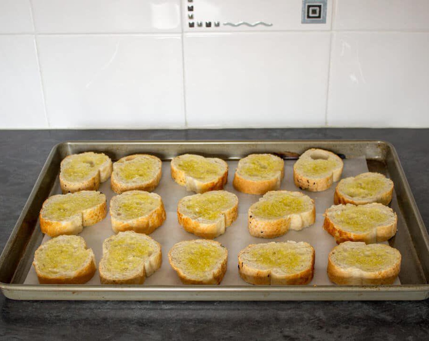 step 2 Slice the Baguette (1) into 1-inch slices then brush one side with Olive Oil (as needed). Place the slices oiled side up onto the prepared baking sheet & pop into the preheated oven for about 10 minutes until golden and crispy.