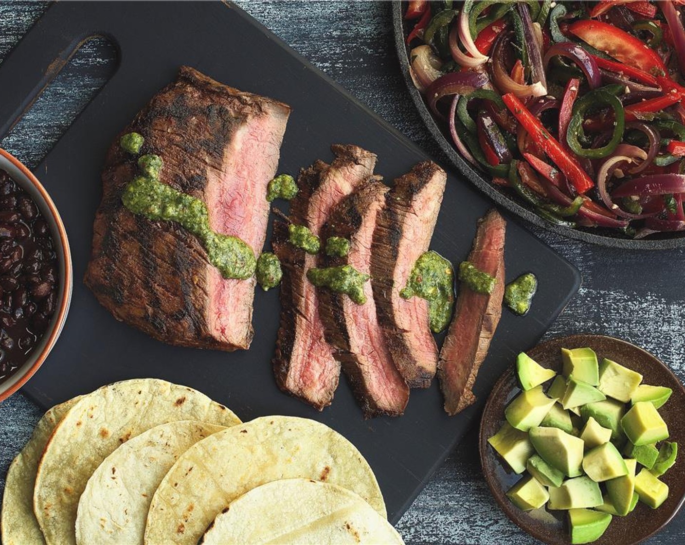step 16 Slice the flank steak in 1/2-inch slices against the grain. Drizzle the chimichurri over the meat. Place bell pepper mixture, cumin lime black beans, avocado, and corn tortillas on the side. Serve family-style.