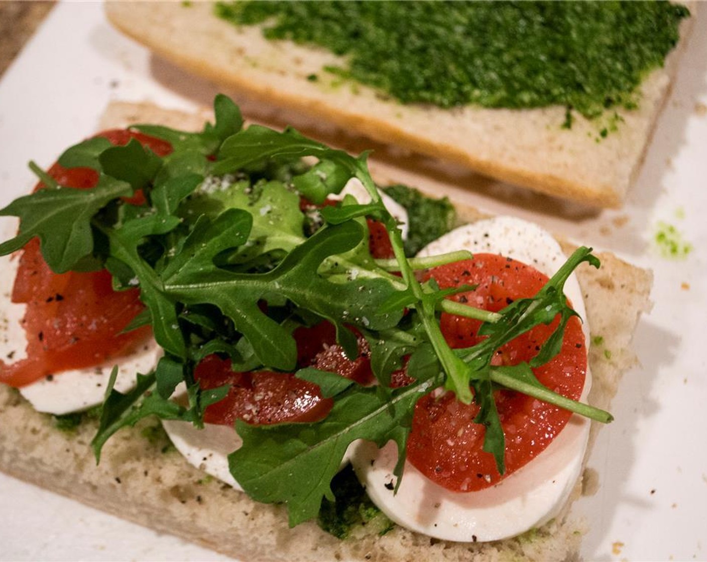 step 5 Oiled side down, liberally spread the pesto on the inside of each half. On the bottom, add the sliced mozzarella, Plum Tomatoes (4), and Arugula (1 bunch). Season with salt and pepper. Place the tops of the bread on each sandwich and place in the Panini Press.