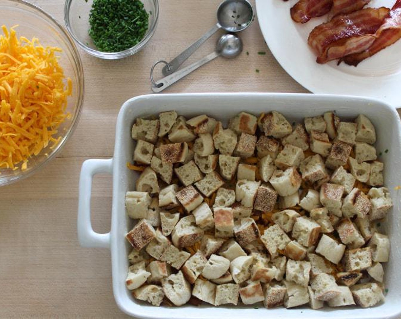 step 7 To assemble, sprinkle half of the bacon, Fresh Chives (1 1/2 Tbsp), and 1 cup of the Sharp Cheddar Cheese (3/4 cup) in that order, onto the bottom of the baking dish. Add the English muffin cubes on top in one even layer.