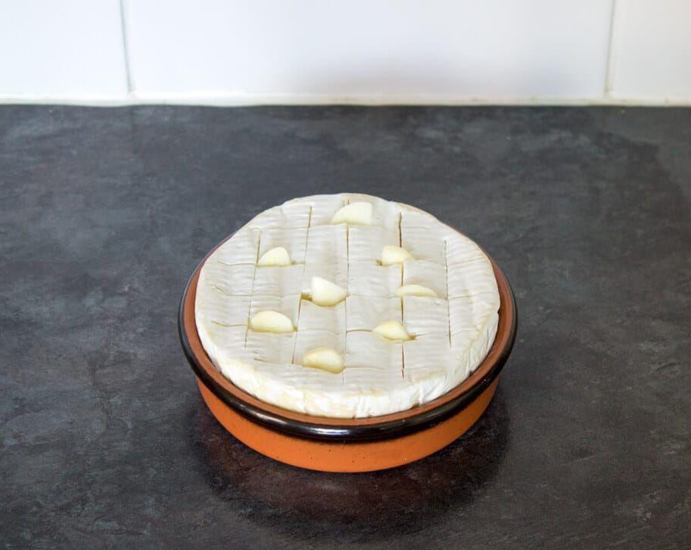 step 2 Remove the Camembert (1 pckg) from it’s plastic wrapper and place either back in its wooden box, or into a fitted dish. Slice a cross-hatch pattern in the top of the cheese. Try not to cut into the sides. Roughly slice the Garlic (1 clove) clove then wedge them into the lines you’ve just cut.