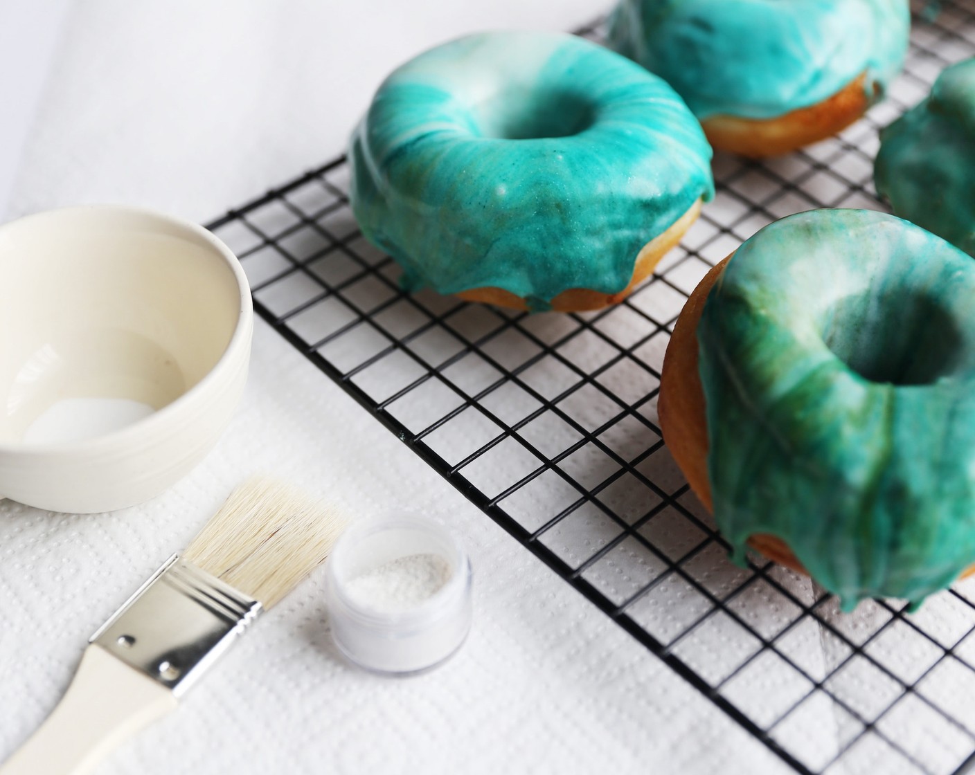 step 22 While the glaze is still wet, add the finishing touches to your Mermaid Doughnuts by using a clean brush to flick additional AmeriColor Soft Gel Paste™ in Bright White (to taste) onto the glaze, and then finishing off with a sprinkle of Edible Glitter (to taste).